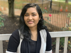 Photo of Adina Shrestha. She has dark, shoulder-length and is wearing a black and white top. She sits on a park bench in a courtyard.