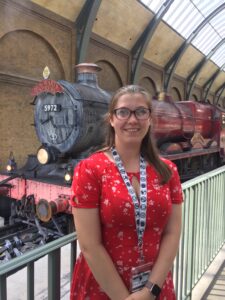 Photo of Anna Zittle. She has long, light-brown hair and is wearing a red sundress. She stands in front of a steam train made to look like the Hogwart's Express from Harry Potter.