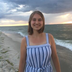 Photo of Allison Berardi. She has short blond hair and is wearing a blue and white striped jumper. She is standing on a beach under cloudy skies at what appears to be sunset.