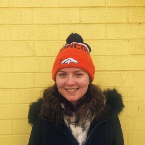 photo of danielle mullan, smiling, wearing winter coat, orange hat, and standing against a yellow brick wall