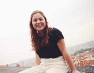 photo of julia dash, smiling; she has long brunette hair wearing white slacks and a black top; she's seated outside on a cloudy day