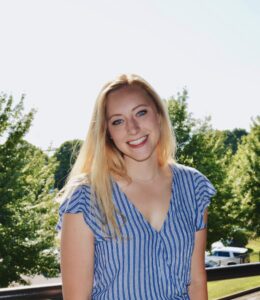 Photo of Maddy Proulx. She has long blond hair and is smiling, her head cocked slightly to one side. She is wearing a blue top and is standing in front of evergreen trees on a sunny day.