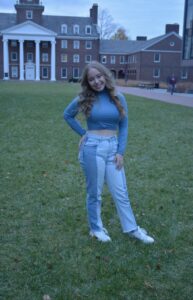 Photo of Lauren. She is wearing jeans and a long sleeve shirt and is standing on grass in front of a building on campus.