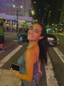 Photo of Emma Chen. She has long brown hair and is smiling. Emma is standing on the street with a car and buildings behind her.