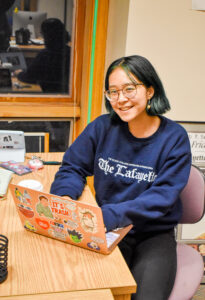 Photo of Shirley Liu. Shirley has short black hair, glasses, and is wearing a blue sweatshirt. Shirley is sitting down at table with a computer.