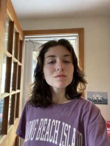 Photo of Emily. She has shoulder length brown hair and is wearing a purple t-shirt. Emily is standing in front of a doorway.