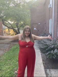 Photo of Lisa. She has long blond hair and is wearing a red jumpsuit. Lisa is standing out a building with her arms spread open.