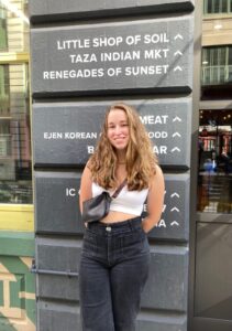 Photo of Tess Stanley. Tess has long blond hair and is wearing a tank top and jeans. She is smiling in front of a sign.