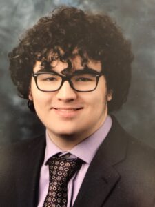 College student with short curly hair wears a suit and tie and smiles at the camera.